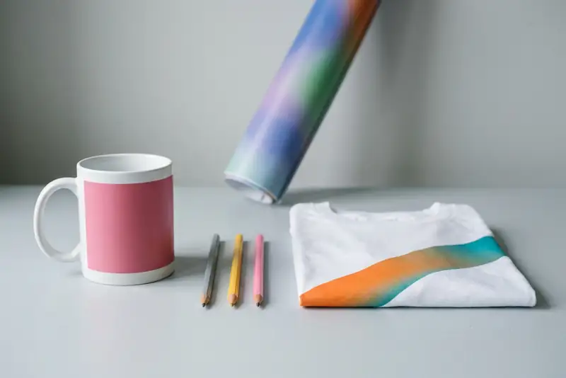 Taza con una banda rosada, tres lápices, una camiseta doblada con una franja de color y un banner enrollado